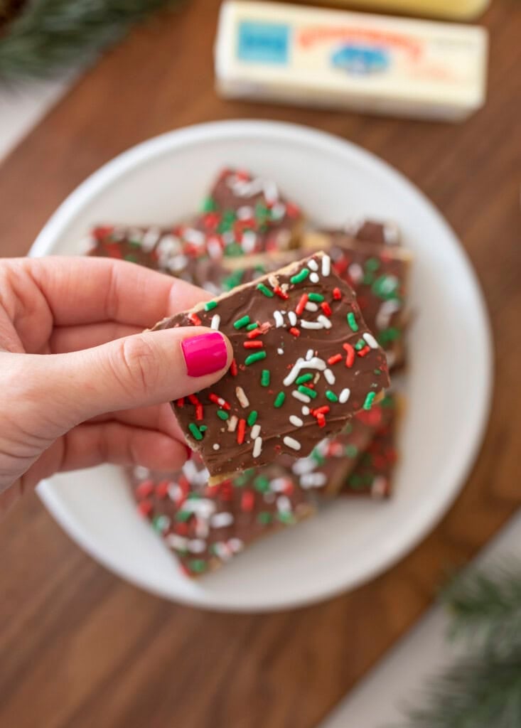 A hand with pink nail polish holds a piece of chocolate bark topped with red, green, and white sprinkles. More pieces are stacked on a white plate in the background.
