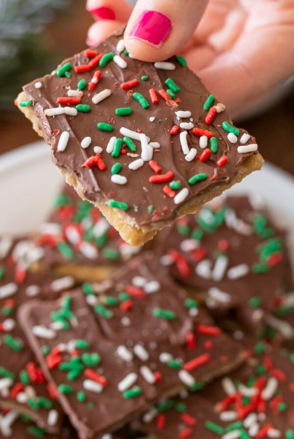 A hand with pink nail polish holds a chocolate-covered cracker topped with red, green, and white sprinkles over a plate of similar treats. Pine branches are blurred in the background.