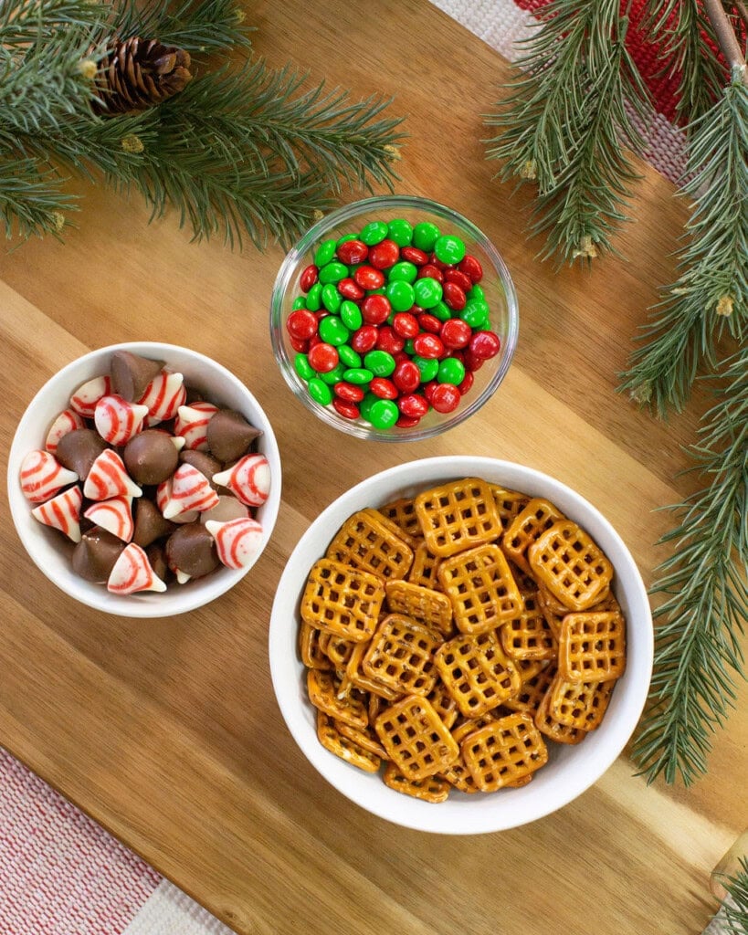Three bowls on a wooden surface: one with red and green candies, one with square pretzels, and one with chocolate and peppermint candies. Pine branches are placed around the bowls for decoration.