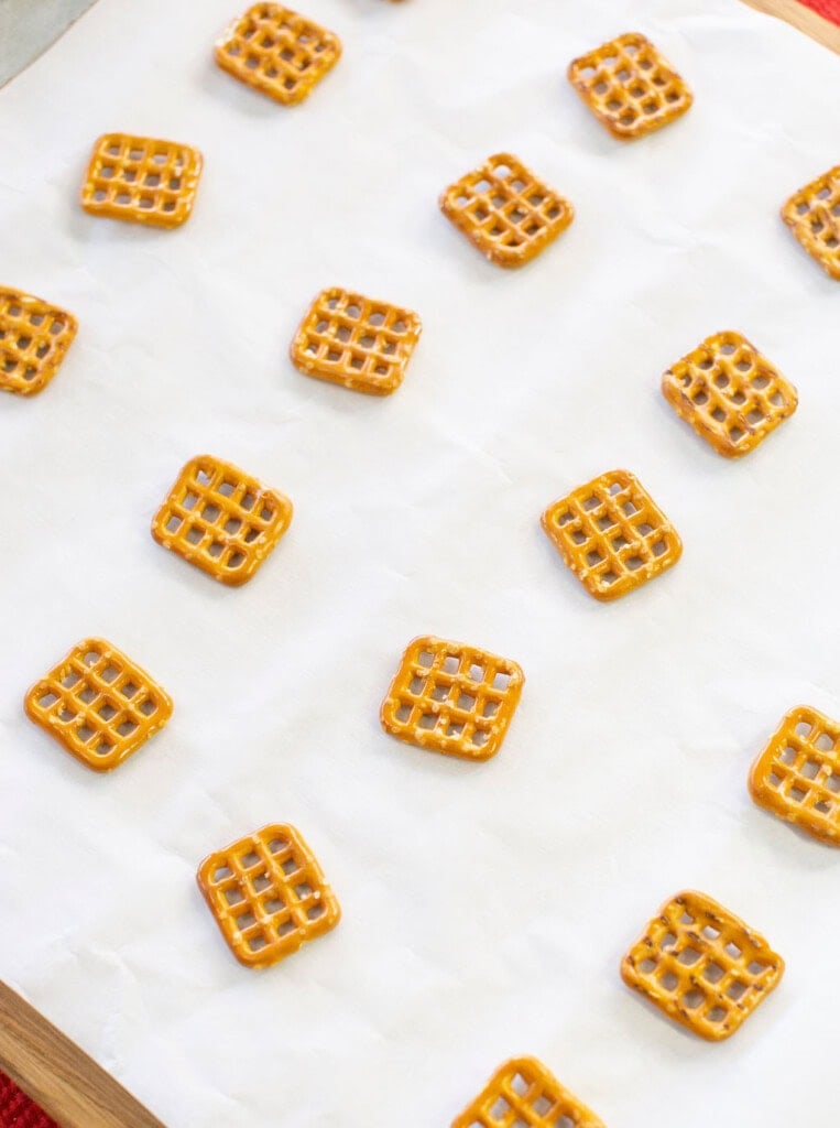 Square pretzels are spaced out on a sheet of white parchment paper, placed on a wooden surface.