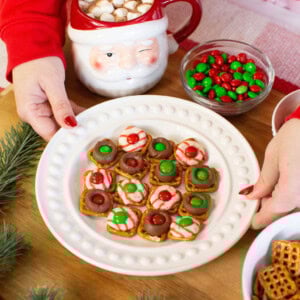 A person in a red sweater holds a plate of holiday pretzel treats topped with chocolate, candy, and festive colors. Nearby are a Santa mug with hot chocolate and marshmallows, a bowl of red and green candies, and some pine branches.