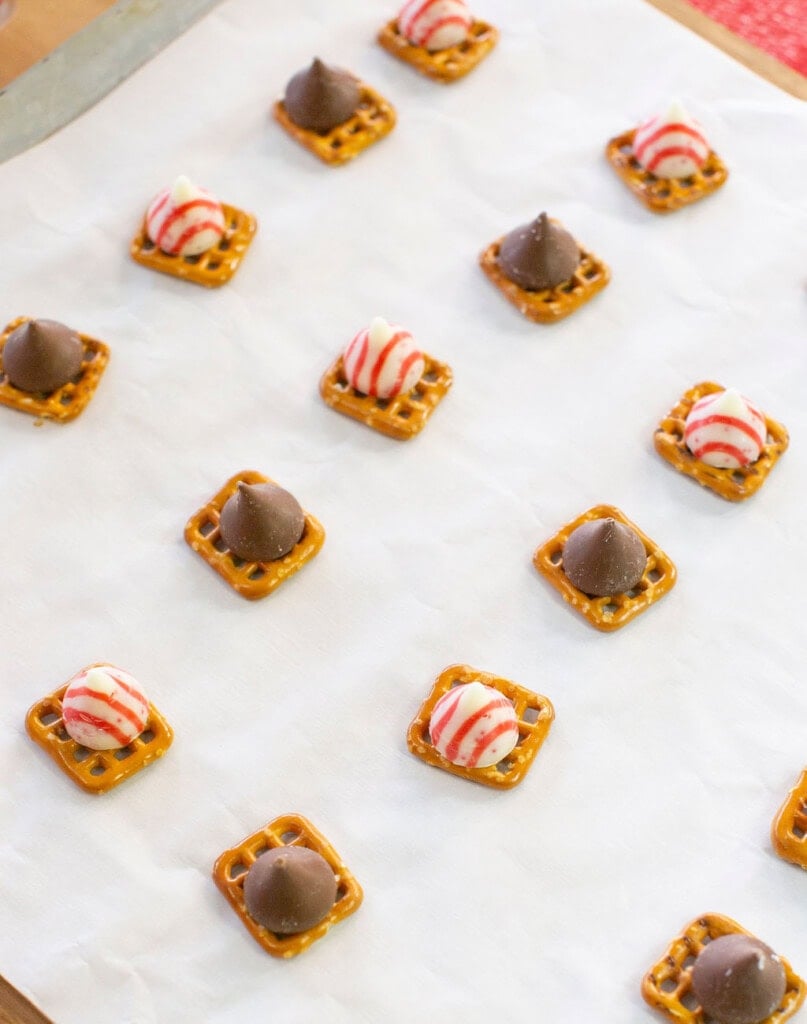 Small square pretzels topped with either striped peppermint or chocolate kisses are arranged on a sheet of parchment paper for baking or cooling.