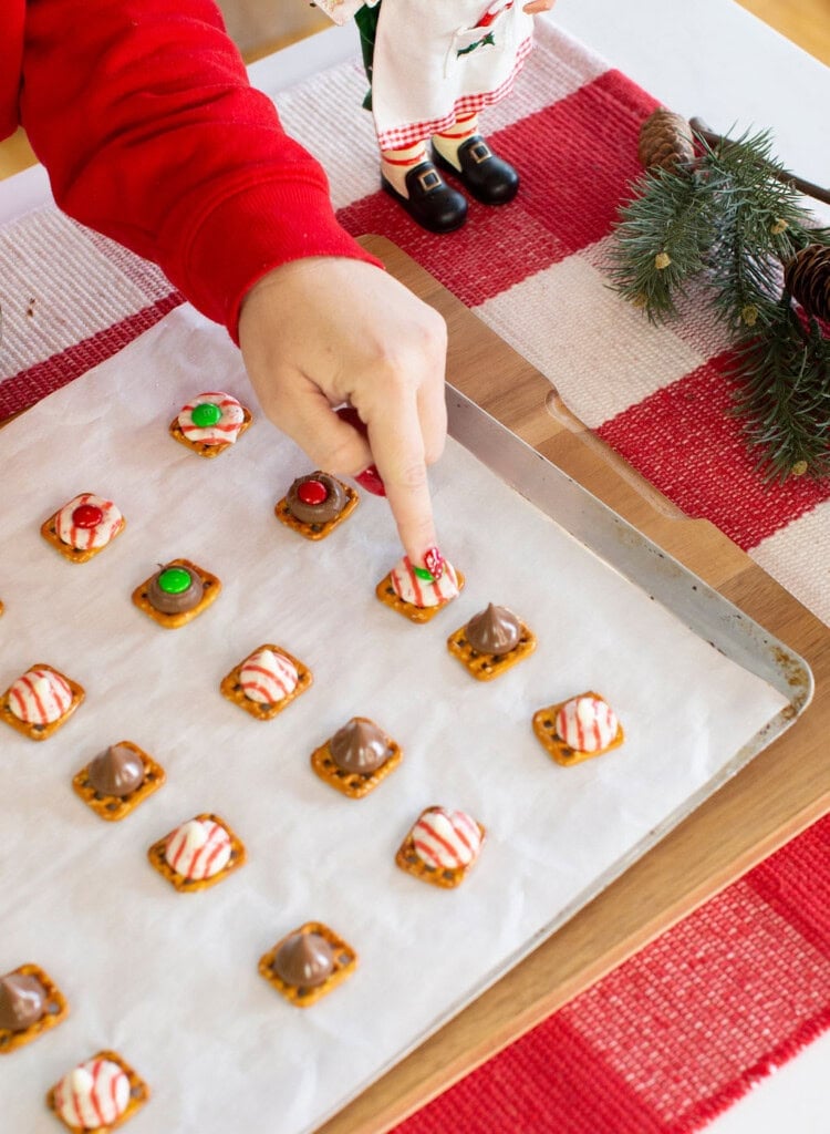 A hand in a red sleeve points to a holiday pretzel treat on a baking sheet lined with parchment paper. The sheet holds various pretzel snacks topped with candy. Festive decor and a red plaid tablecloth are visible nearby.
