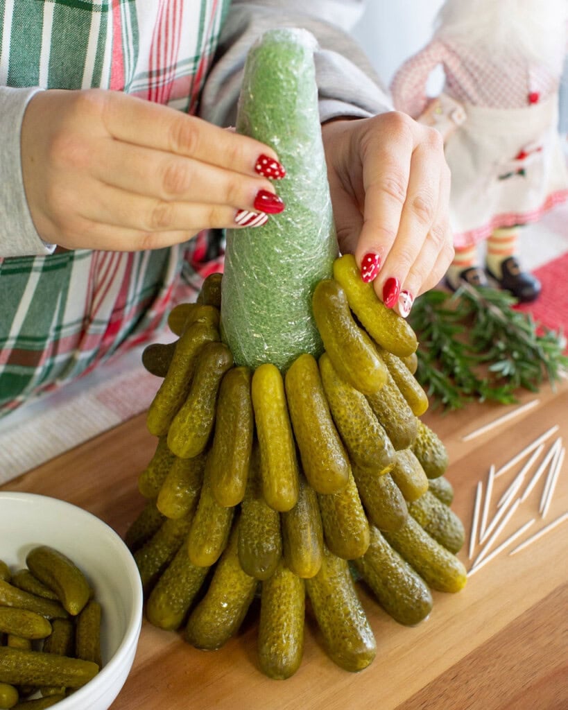 A person with festive, red-striped nails arranges pickles around a cone-shaped base to create a pickle Christmas tree. A bowl of pickles, rosemary, toothpicks, and holiday decor are nearby on a wooden surface.