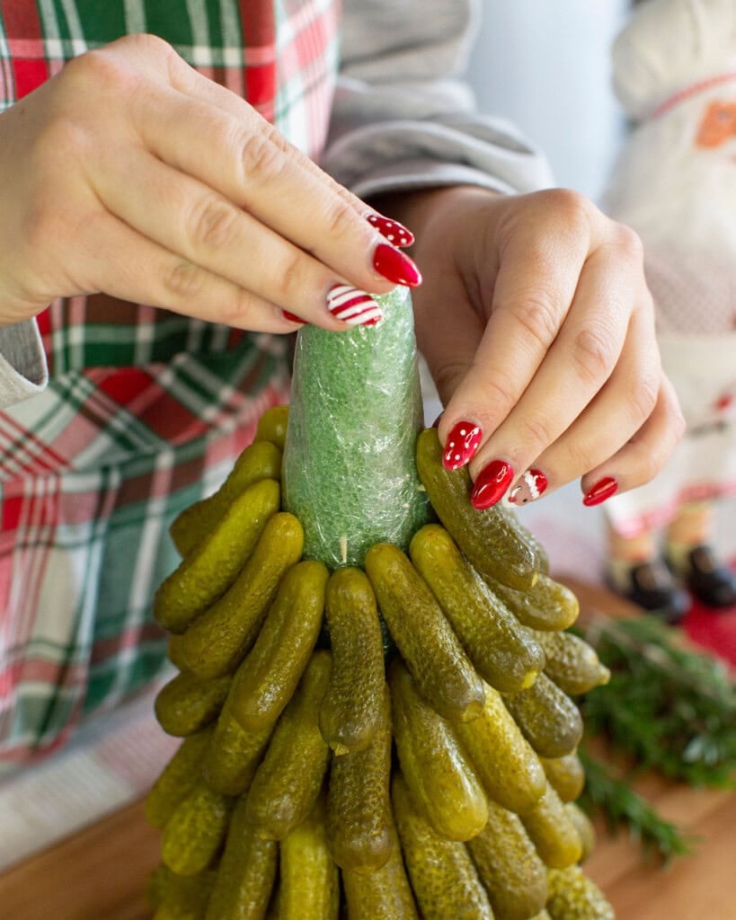 A person with festive red and white nail art arranges pickles on a green cone, creating a Christmas tree-shaped holiday centerpiece. The person is wearing a plaid apron.