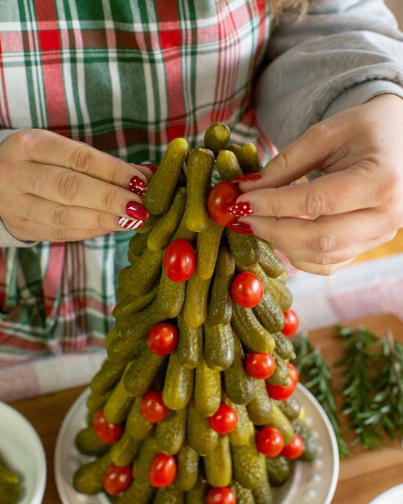 A person with festive, red-painted nails arranges grape tomatoes on a Christmas tree-shaped arrangement of pickles. They are wearing a plaid apron, and rosemary sprigs are visible nearby.