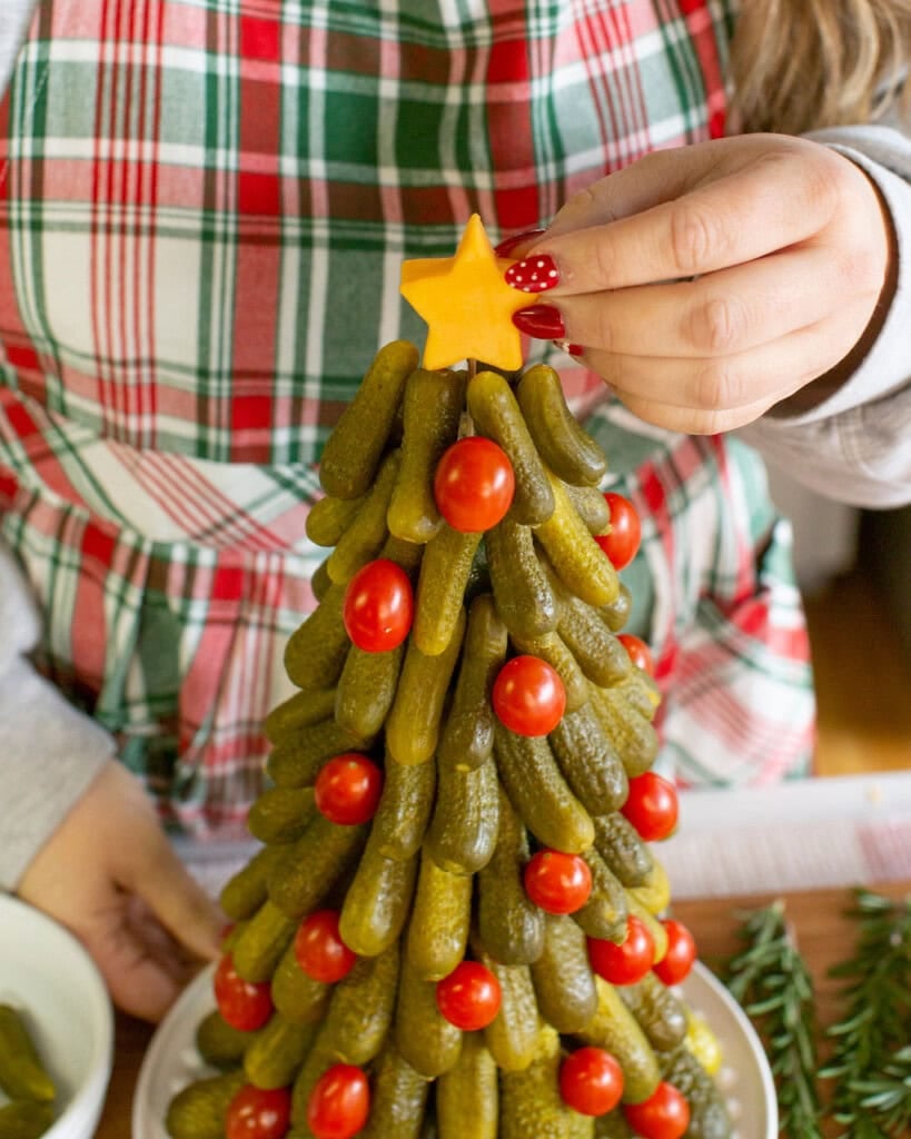 A person in a plaid apron places a yellow cheese star on top of a Christmas tree-shaped arrangement made of pickles and cherry tomatoes.