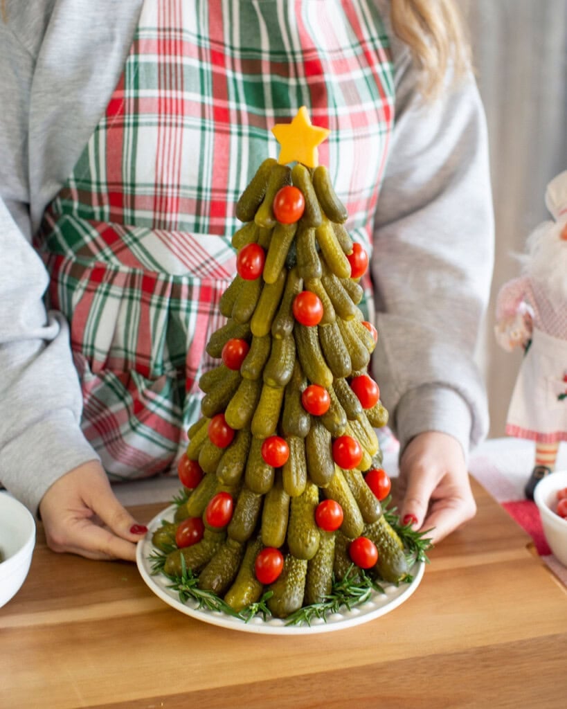 A person in a plaid apron holds a plate with a Christmas tree-shaped arrangement of pickles and cherry tomatoes, topped with a yellow star and surrounded by sprigs of rosemary.
