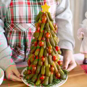 A person in a plaid apron holds a platter with a Christmas tree made of stacked pickles and cherry tomatoes, topped with a cheese star and garnished with rosemary.