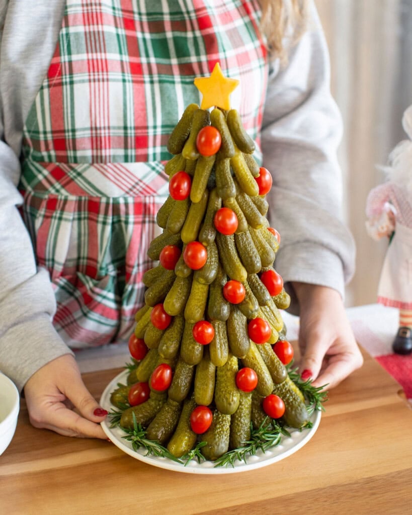 A person in a plaid apron holds a platter with a Christmas tree made of stacked pickles and cherry tomatoes, topped with a cheese star and garnished with rosemary.