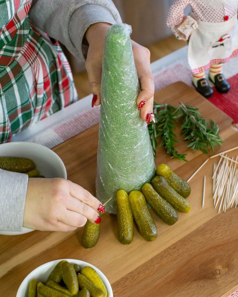 A person wraps a green foam cone in plastic wrap and arranges pickles upright around the base, preparing a food display on a wooden table. Bowls of pickles, rosemary, and toothpicks are nearby.
