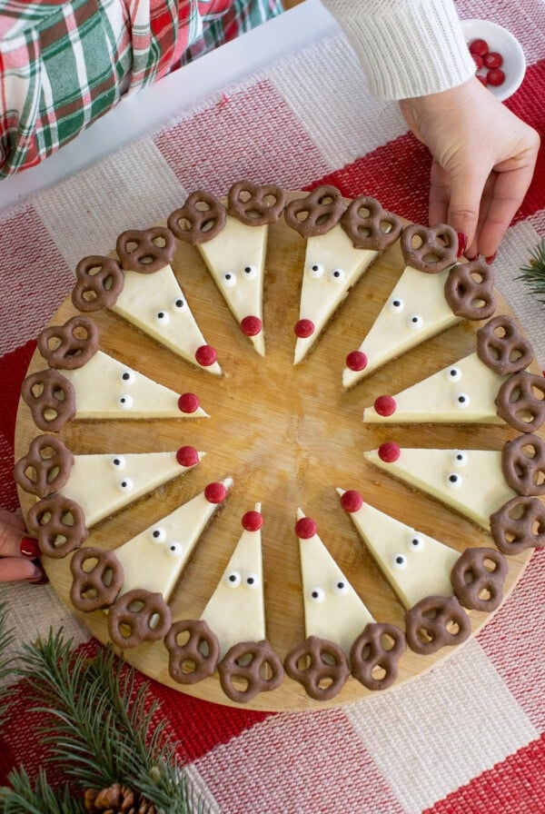 Cheesecake slices decorated as reindeer with pretzel antlers, candy eyes, and red candy noses, arranged in a circle on a wooden board. Hands are placing the board on a red and white tablecloth.