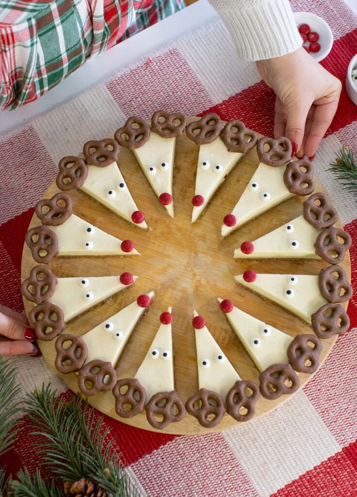 Cheesecake slices decorated as reindeer with pretzel antlers, candy eyes, and red candy noses, arranged in a circle on a wooden board. Hands are placing the board on a red and white tablecloth.