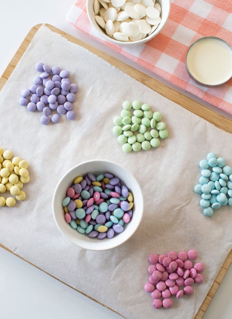 Six small piles of colorful candy-coated chocolates are arranged on parchment paper, with a bowl of mixed candies in the center. Nearby are a bowl of white chocolate wafers and a cup of liquid on a pink-checked cloth.