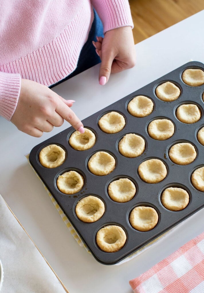 A person in a pink sweater touches a mini tart shell in a muffin tin filled with empty, baked tart shells on a white countertop.