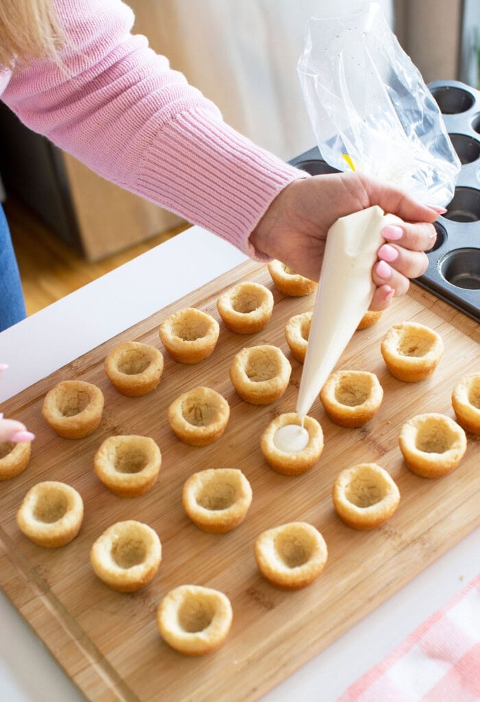 A person uses a piping bag to fill hollowed-out cupcake bites arranged on a wooden board; a muffin tin is visible nearby.