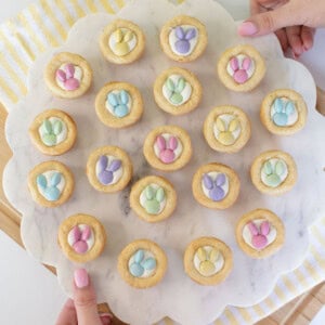 A person holds a round marble tray with 19 cookies topped with pastel-colored candy decorations shaped like bunny paw prints. The tray is on a striped yellow and white cloth over a wooden board.