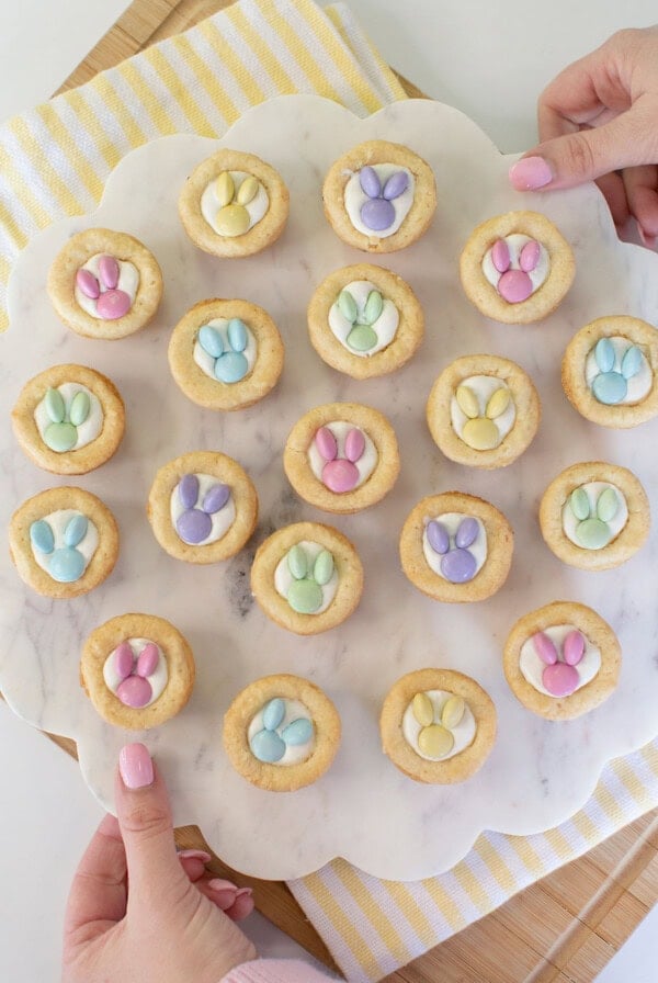 A person holds a round marble tray with 19 cookies topped with pastel-colored candy decorations shaped like bunny paw prints. The tray is on a striped yellow and white cloth over a wooden board.