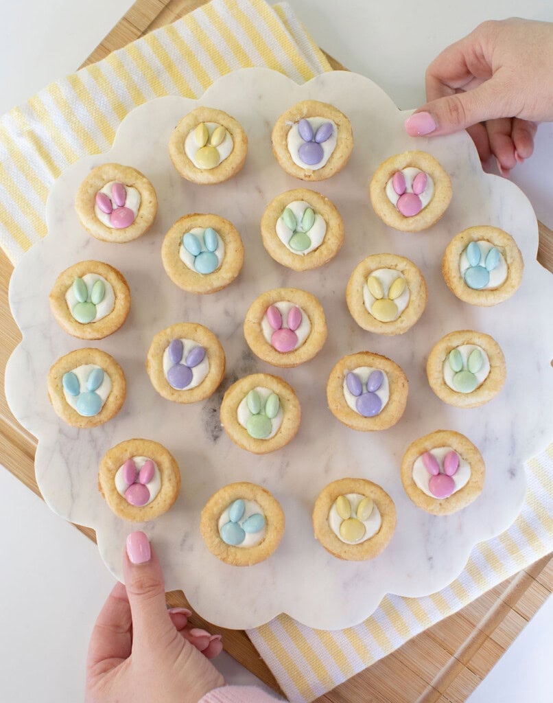 A person holds a round marble tray with 19 cookies topped with pastel-colored candy decorations shaped like bunny paw prints. The tray is on a striped yellow and white cloth over a wooden board.