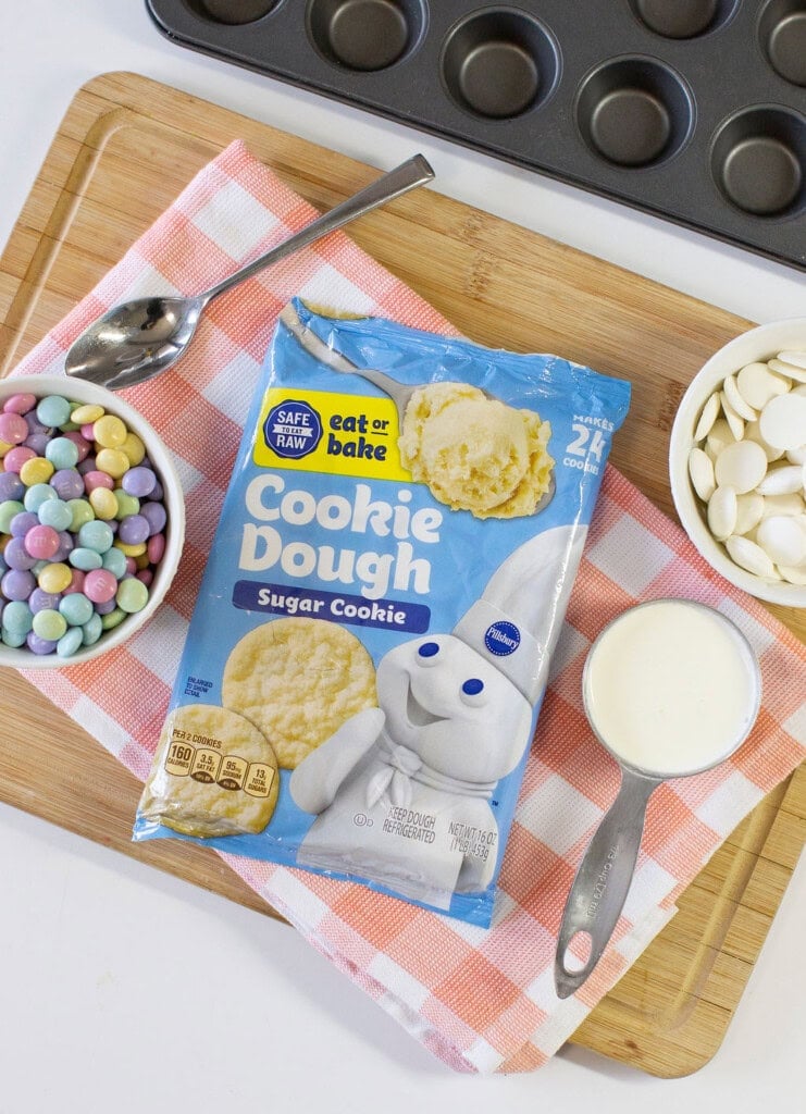 A bag of Pillsbury sugar cookie dough is on a wooden board with a red-and-white checkered towel, surrounded by pastel chocolate candies, white chocolate wafers, and a bowl of milk near a muffin pan.