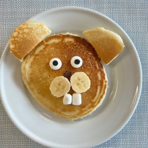 A pancake arranged on a plate to look like a dog’s face, with banana slices for cheeks, marshmallows for teeth, chocolate chips for a nose, candy eyes, and pancake pieces for ears.