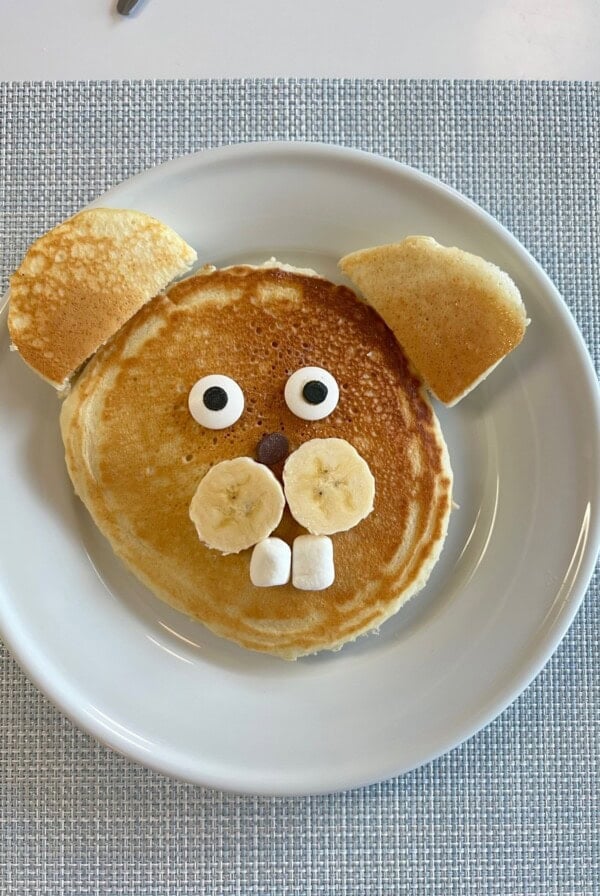 A pancake arranged on a plate to look like a dog’s face, with banana slices for cheeks, marshmallows for teeth, chocolate chips for a nose, candy eyes, and pancake pieces for ears.