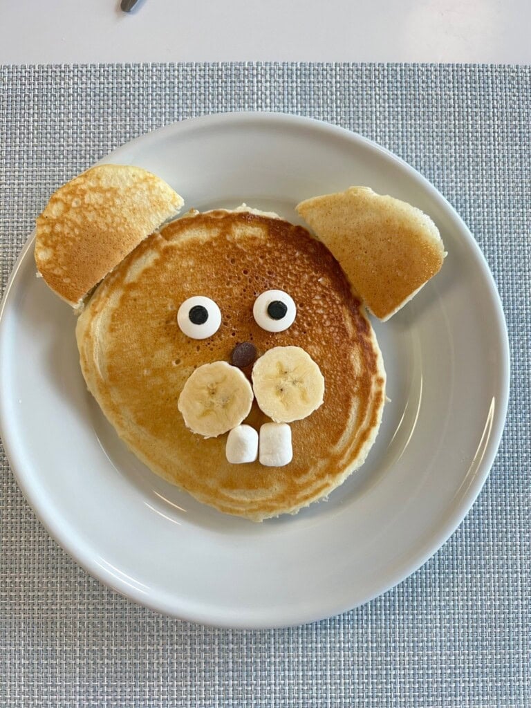 A pancake arranged on a plate to look like a dog’s face, with banana slices for cheeks, marshmallows for teeth, chocolate chips for a nose, candy eyes, and pancake pieces for ears.