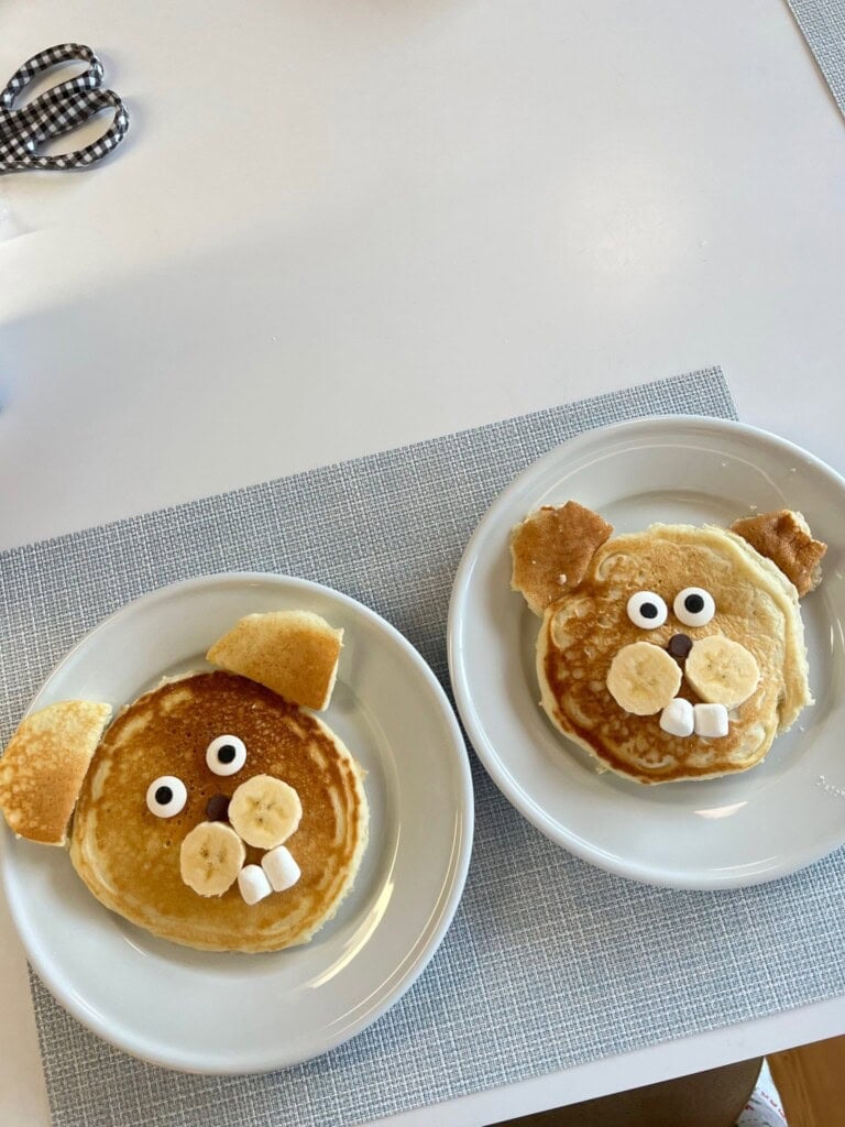 Two plates each with a pancake decorated to look like an animal face using banana slices for cheeks, chocolate chips for eyes, marshmallows for teeth, and pancake pieces for ears.