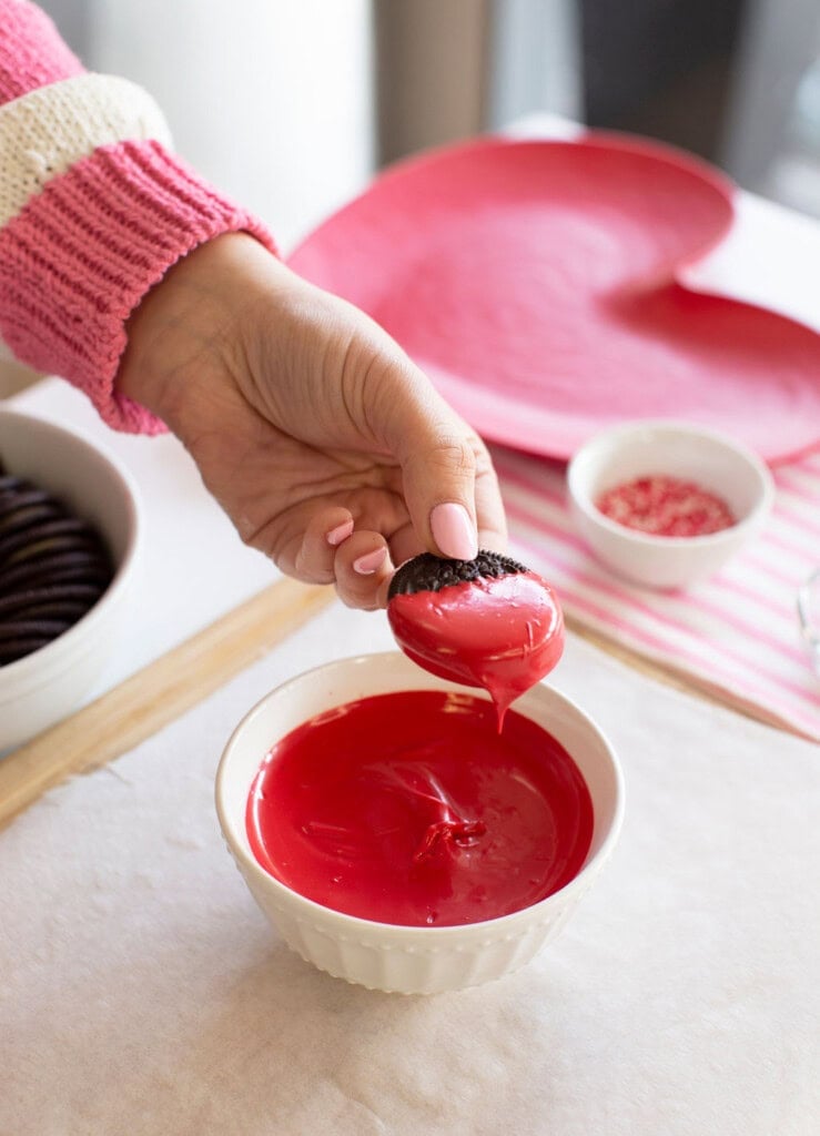A hand in a pink sweater dips a cookie halfway into a bowl of red melted chocolate, with a heart-shaped plate and bowls of cookies and sprinkles in the background.