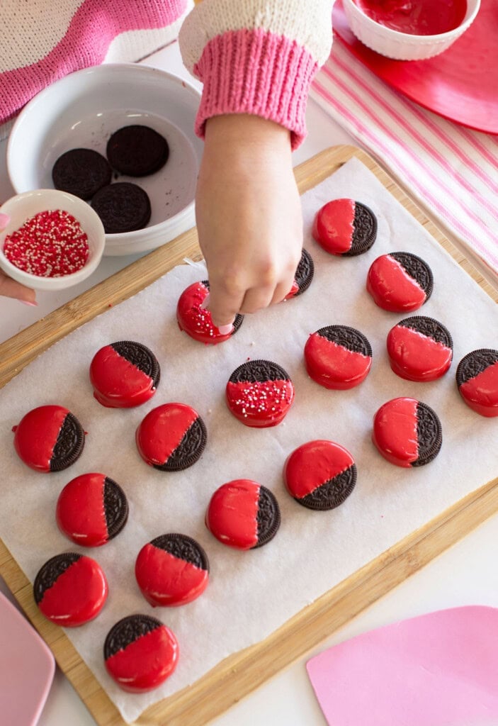 A person sprinkles red and white sprinkles onto half-dipped red chocolate Oreo cookies arranged on parchment paper, with bowls of sprinkles and cookies nearby on a wooden board.