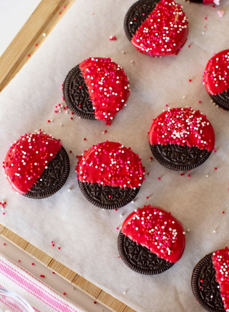 Chocolate sandwich cookies half-dipped in bright red icing and decorated with red, white, and pink sprinkles, arranged on parchment paper.