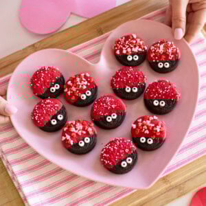A pink heart-shaped plate holds Oreo cookies decorated with red icing, candy eyes, and heart-shaped sprinkles. The plate is on a pink-striped cloth, and hands are holding the plate.