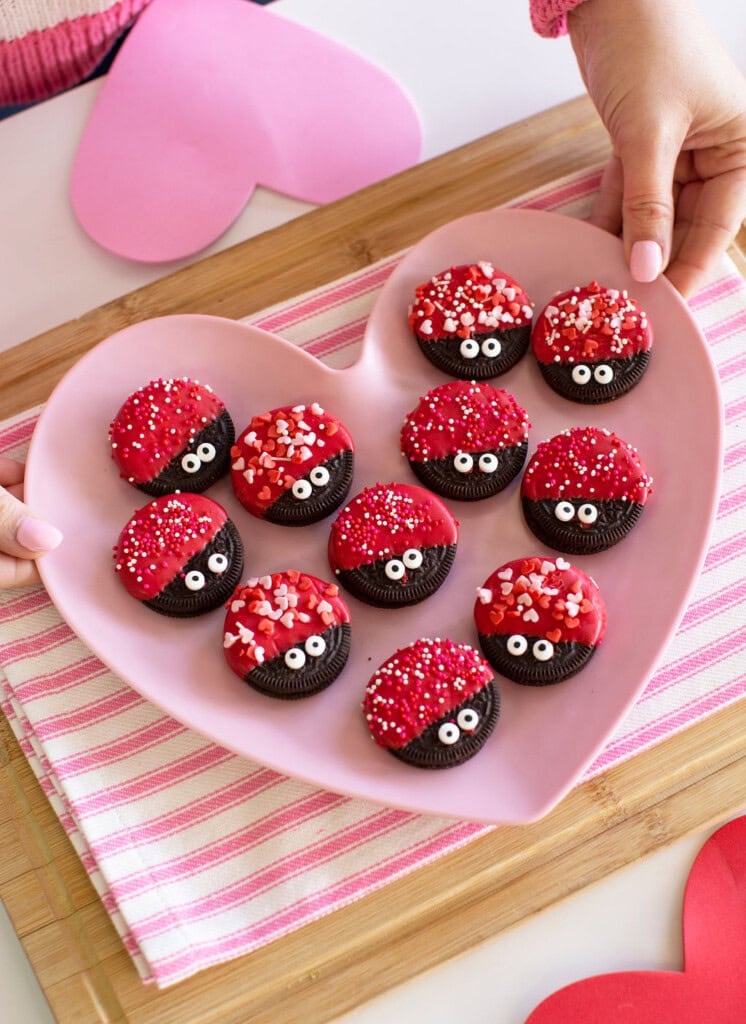 A pink heart-shaped plate holds Oreo cookies decorated with red icing, candy eyes, and heart-shaped sprinkles. The plate is on a pink-striped cloth, and hands are holding the plate.