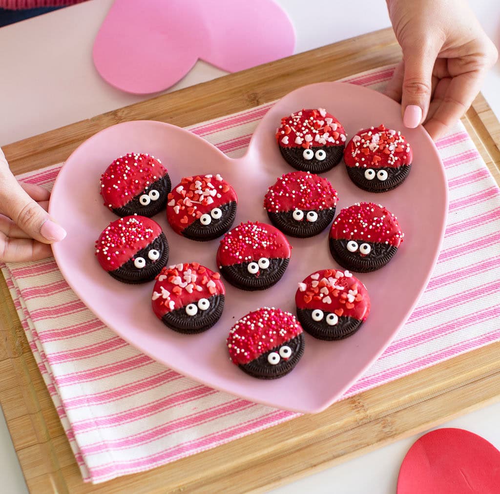 A pink heart-shaped plate holds Oreo cookies decorated with red frosting, sprinkles, and candy eyes to look like ladybugs. The plate is on a striped cloth, and hands are holding the plate. Paper hearts are nearby.