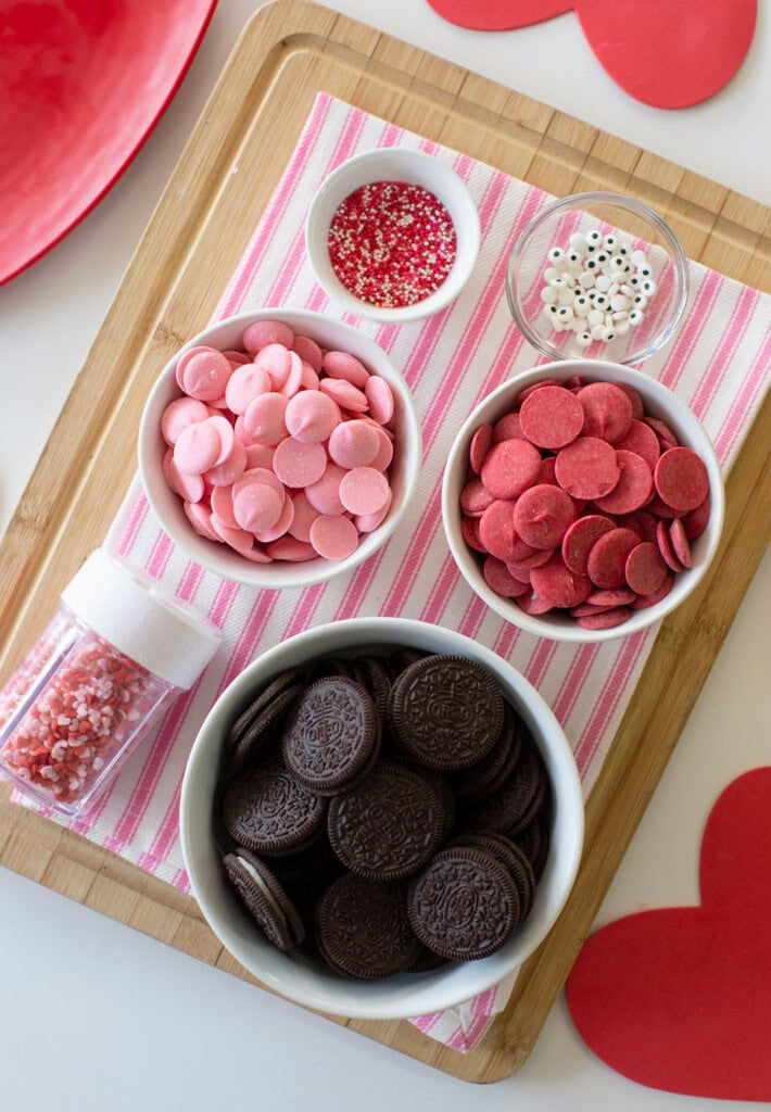 A wooden board holds bowls of chocolate sandwich cookies, pink and red candy melts, and small containers of heart and round sprinkles, all arranged on a pink-striped cloth with red paper hearts nearby.