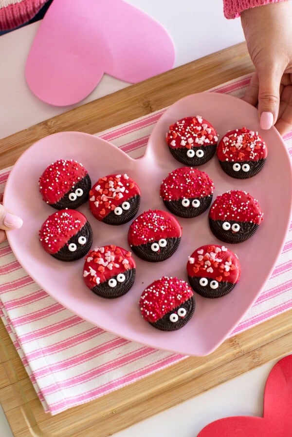 A heart-shaped pink plate holds chocolate cookies dipped in red coating, decorated with candy eyes and heart-shaped sprinkles. Hands are holding the plate over a pink striped cloth and heart-shaped paper cutouts.