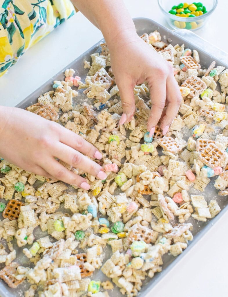 A person arranges a cereal snack mix with colorful candies, pretzels, and assorted cereals on a baking sheet. A small bowl of additional candies sits nearby on a white surface.