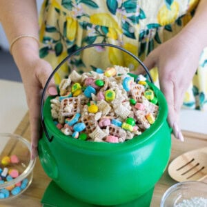A person in a yellow, lemon-patterned dress holds a green plastic cauldron filled with Chex cereal and colorful marshmallows, with a small bowl of marshmallows and a bowl of powdered sugar nearby.