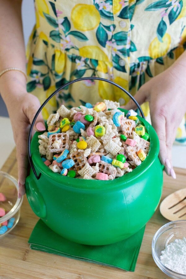 A person in a yellow, lemon-patterned dress holds a green plastic cauldron filled with Chex cereal and colorful marshmallows, with a small bowl of marshmallows and a bowl of powdered sugar nearby.