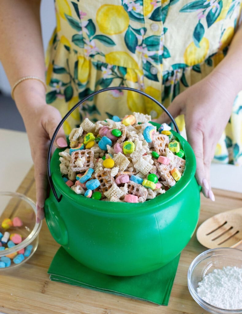 A person in a yellow, lemon-patterned dress holds a green plastic cauldron filled with Chex cereal and colorful marshmallows, with a small bowl of marshmallows and a bowl of powdered sugar nearby.