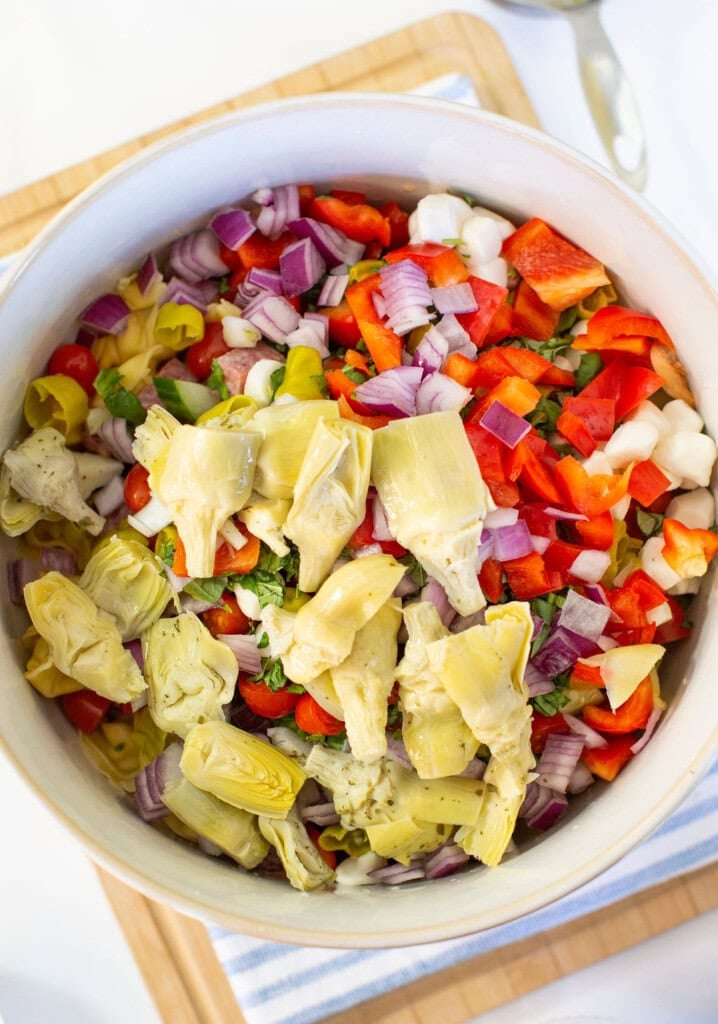 A bowl of colorful chopped vegetables including red onion, red bell pepper, artichoke hearts, mozzarella balls, and fresh herbs arranged on a wooden board.