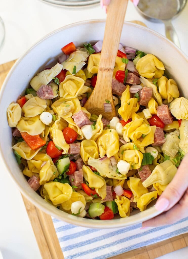 A hand stirs a bowl of pasta salad with tortellini, diced salami, mozzarella balls, red bell peppers, cucumbers, and artichoke hearts using a wooden spoon. The bowl sits on a wooden surface.