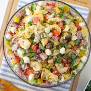 A glass bowl filled with pasta salad containing tortellini, diced salami, mozzarella balls, chopped red peppers, red onions, pepperoncini, and fresh basil, placed on a striped cloth on a wooden board.