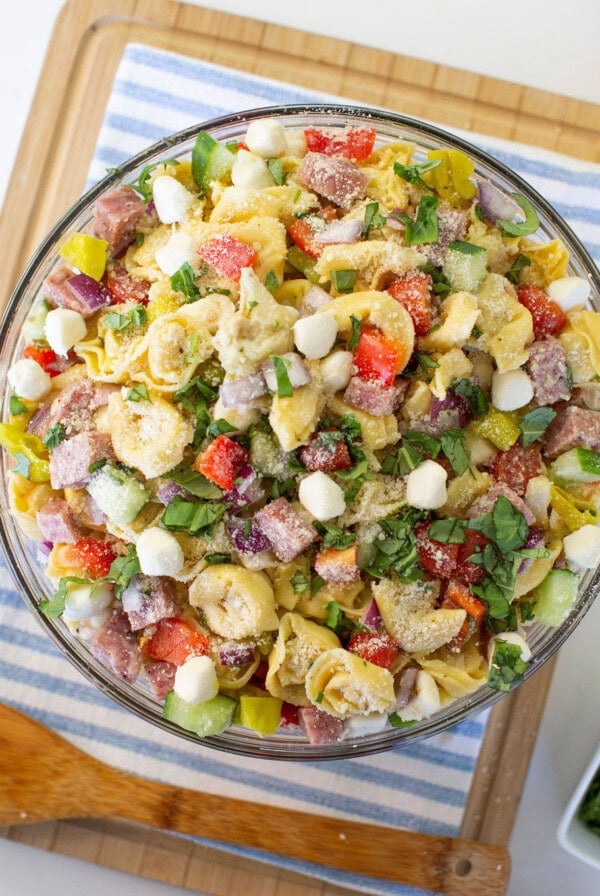 A glass bowl filled with pasta salad containing tortellini, diced salami, mozzarella balls, chopped red peppers, red onions, pepperoncini, and fresh basil, placed on a striped cloth on a wooden board.