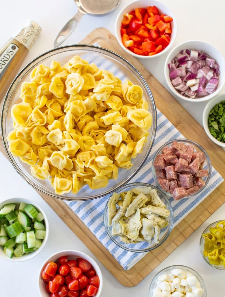 A bowl of uncooked tortellini surrounded by bowls of chopped red bell pepper, red onion, cucumber, salami, artichoke hearts, cherry tomatoes, mozzarella balls, and fresh herbs on a striped cloth and wooden board.