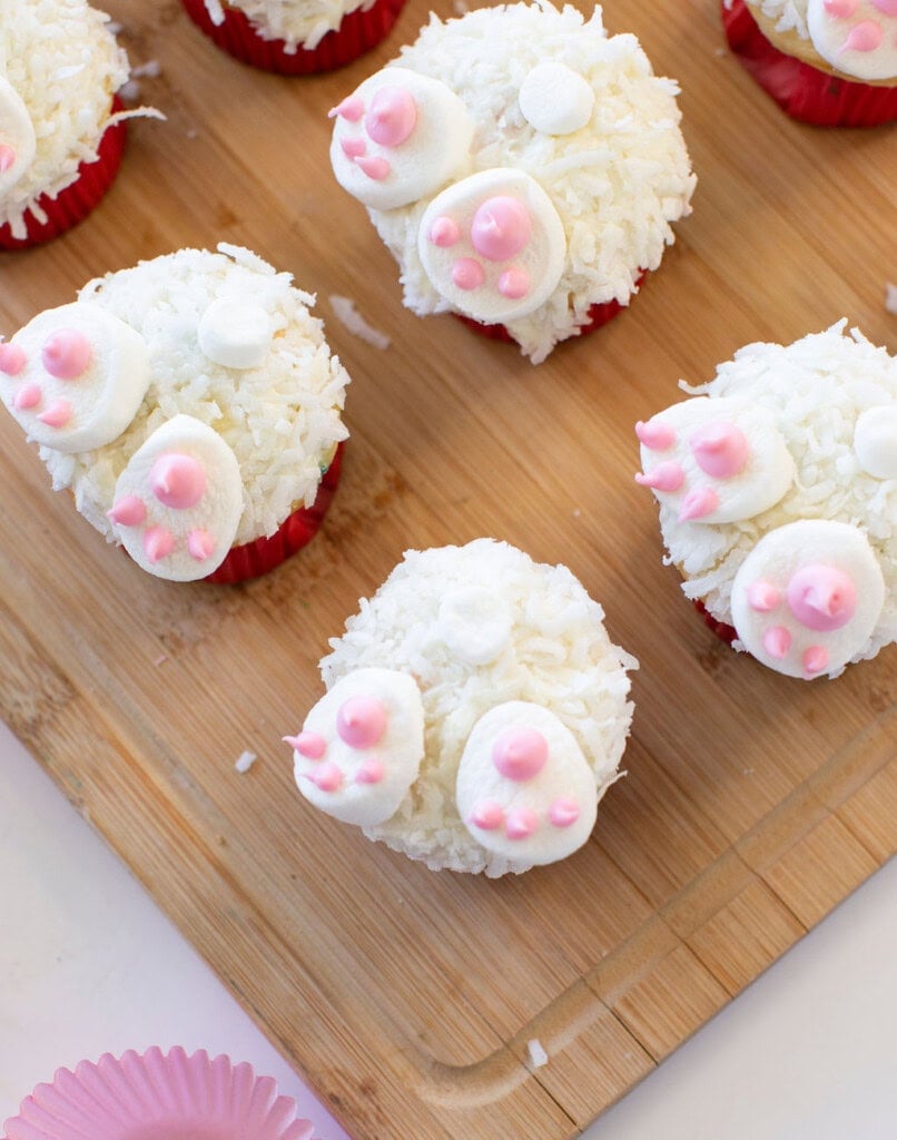 Cupcakes decorated to look like bunny butts, topped with white shredded coconut, marshmallows for feet and tails, and pink icing details, arranged on a wooden board.