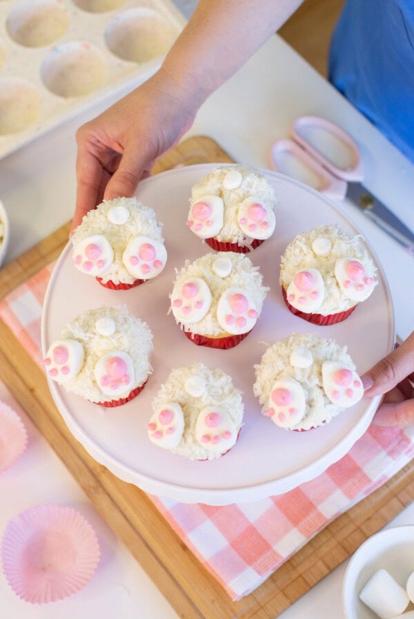 A person holds a white cake stand with six cupcakes decorated to look like bunny butts, featuring white icing, shredded coconut, and pink paw prints. Pink cupcake liners and baking tools are visible nearby.