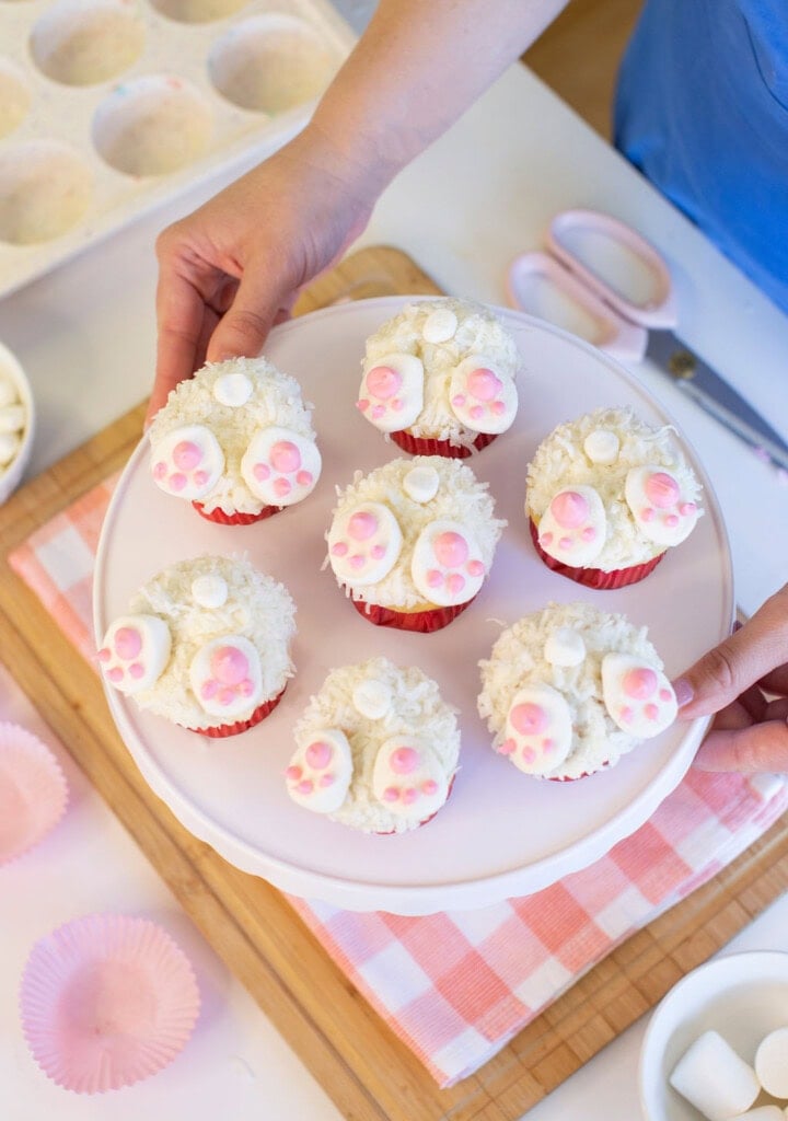 A person holds a white cake stand with six cupcakes decorated to look like bunny butts, featuring white icing, shredded coconut, and pink paw prints. Pink cupcake liners and baking tools are visible nearby.