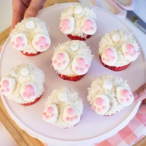 A plate of seven cupcakes decorated to look like bunny bottoms, with white frosting, coconut flakes, and pink paw prints made from icing or fondant. Hands are holding the plate over a table.