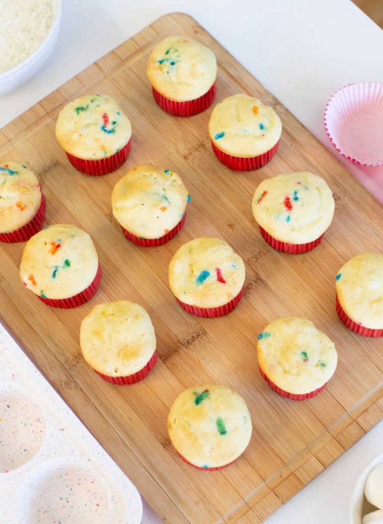 Twelve funfetti cupcakes with red liners are arranged on a wooden cutting board, surrounded by baking supplies, including a white bowl and pink cupcake liners.